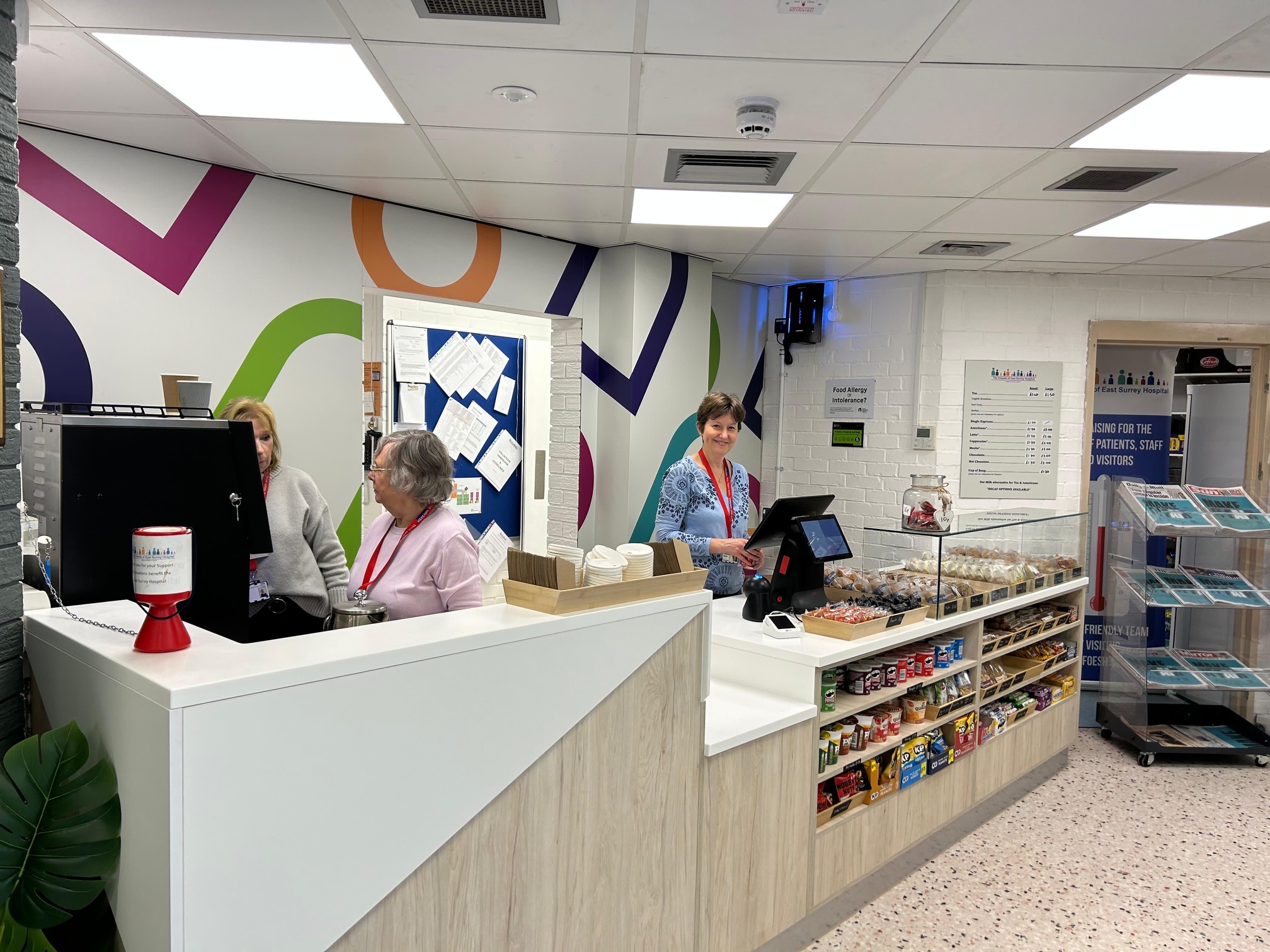 Three women standing behind the coffee shop counter and smiling at the camera. The shop is stocked with chocolate, snacks and refrigerated drinks